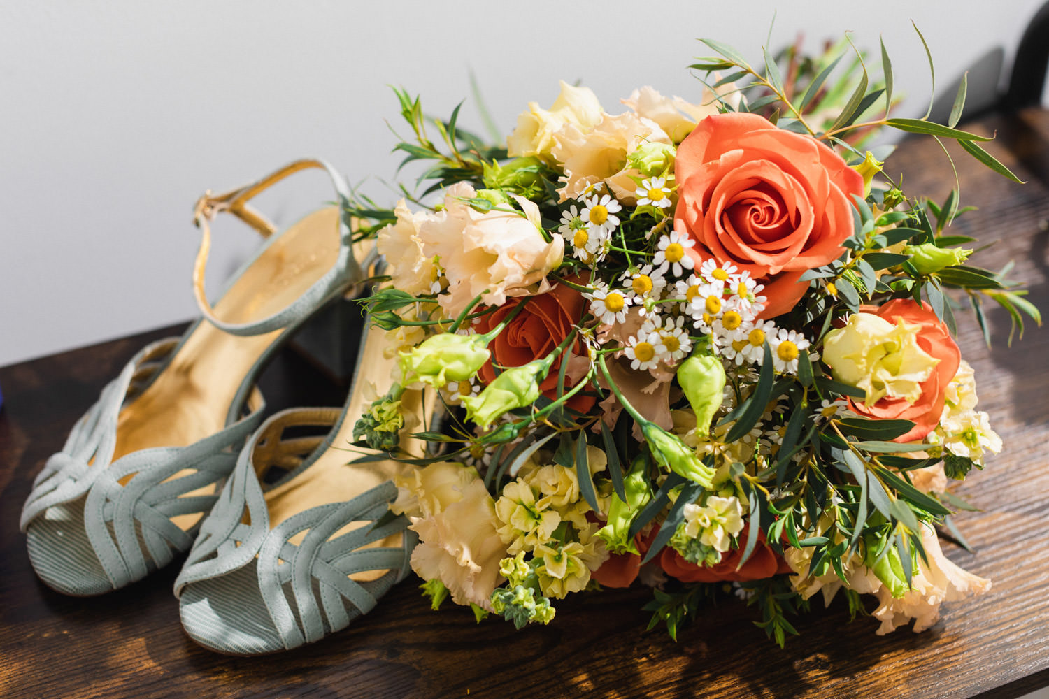 boda de otoño en el Castillo de la Arguijuela 2 Detalle de ramo de novia con rosas naranjas y margaritas junto a sandalias verde menta sobre mesa de madera.