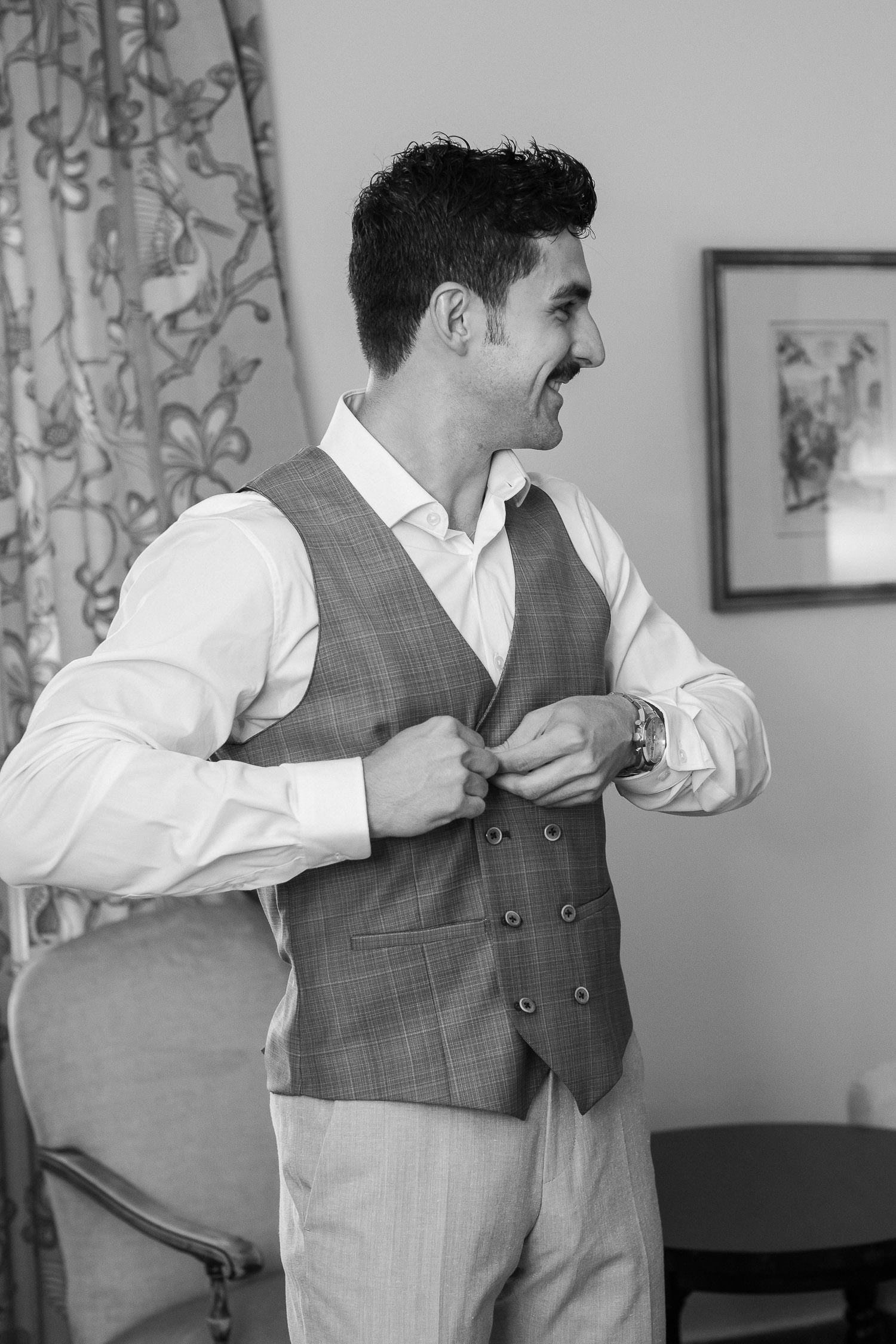 Una boda civil en el Parador de Jarandilla 1 Un novio sonriente, con bigote y peinado impecable, se abrocha un chaleco de traje a cuadros en una elegante habitación con cortinas de flores y cuadros antiguos, en los preparativos previos a su boda en el Parador de Jarandilla.