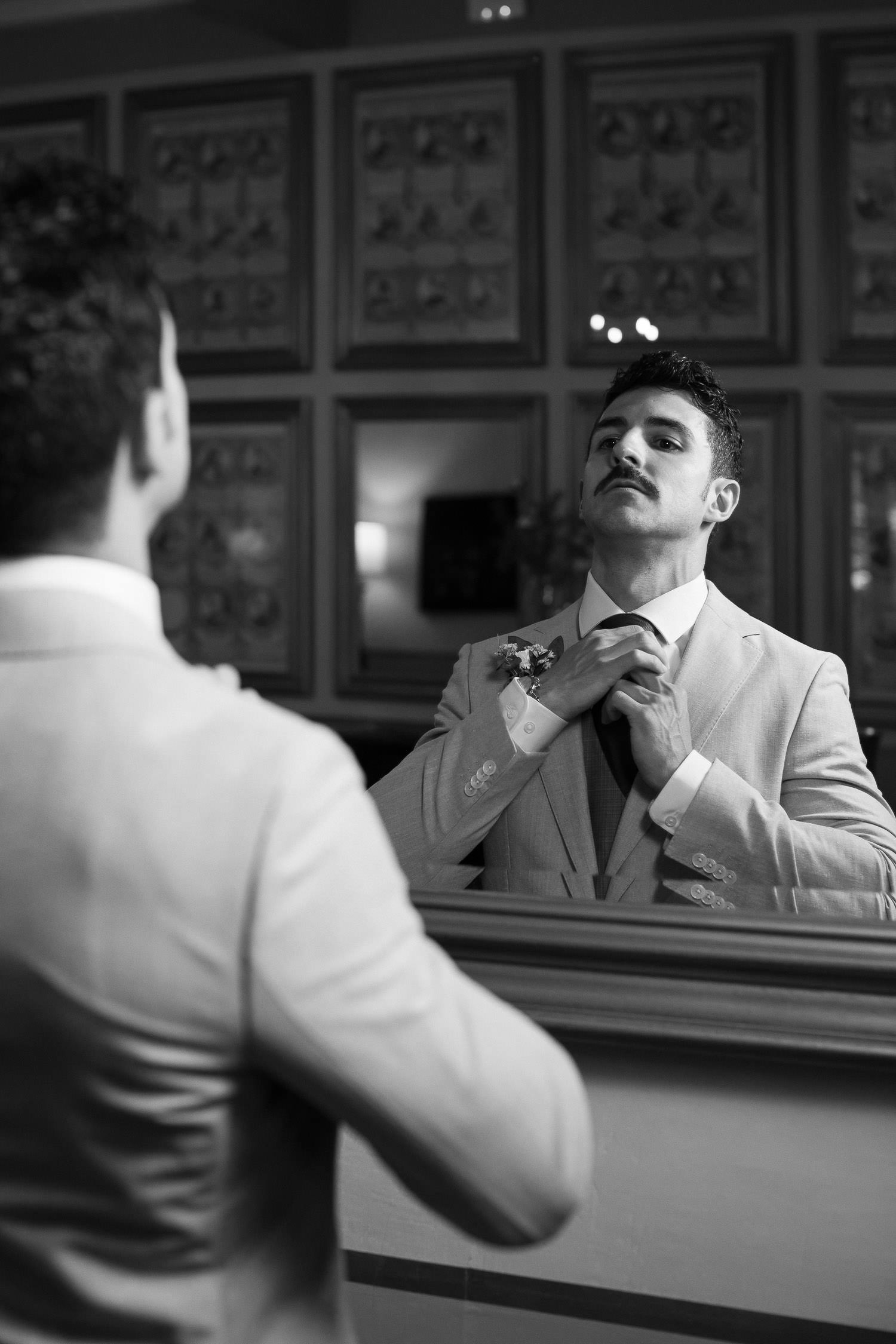 Una boda civil en el Parador de Jarandilla 8 El novio, vestido con un traje claro y con un boutonniere en la solapa, se ajusta la corbata frente al espejo, concentrado en su reflejo mientras se prepara para su gran día.