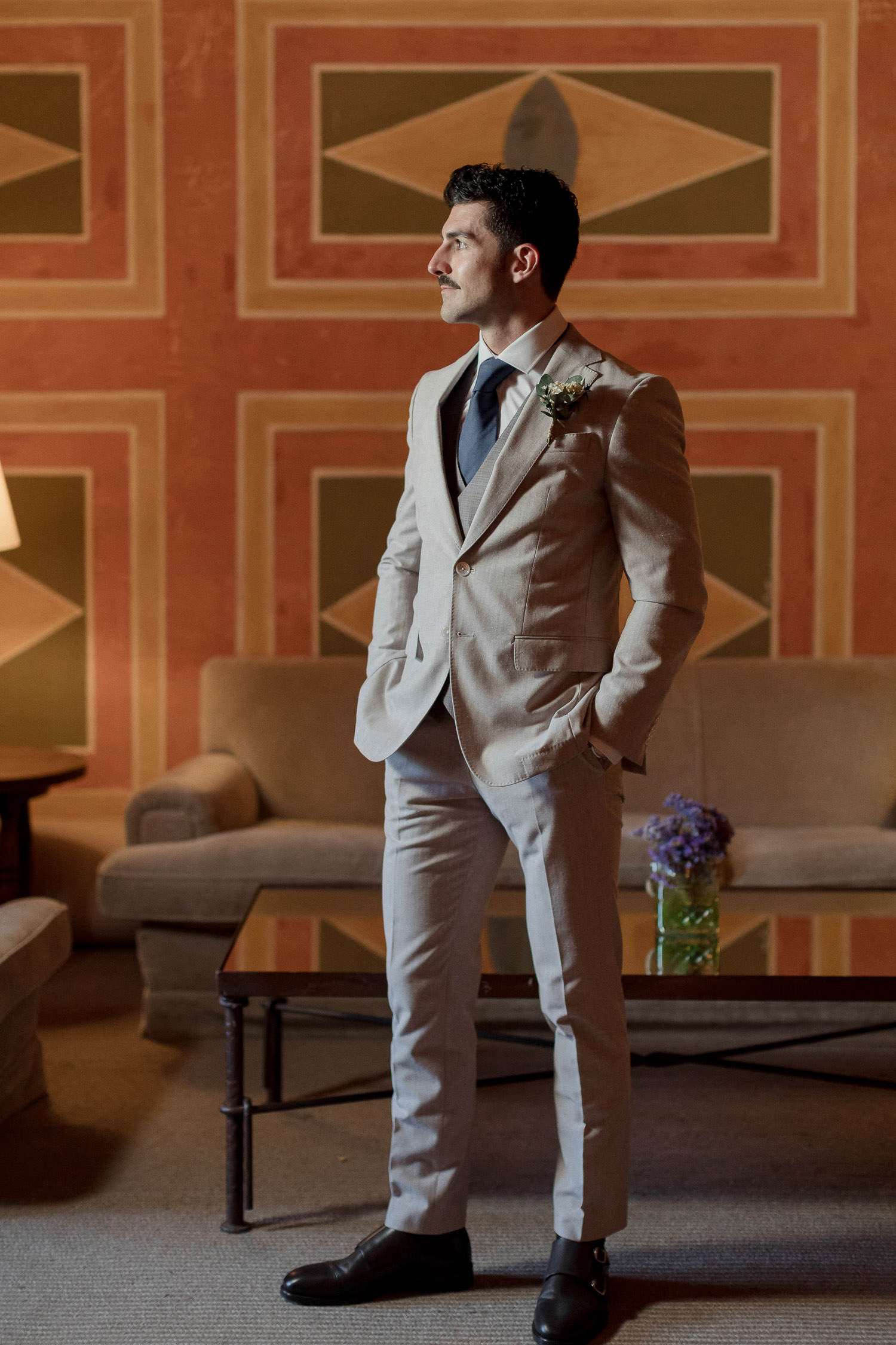 Una boda civil en el Parador de Jarandilla 9 El novio posa de pie, elegante y seguro, con su traje beige y corbata azul marino, en un salón con paredes color terracota decoradas con motivos geométricos, creando un ambiente sofisticado.