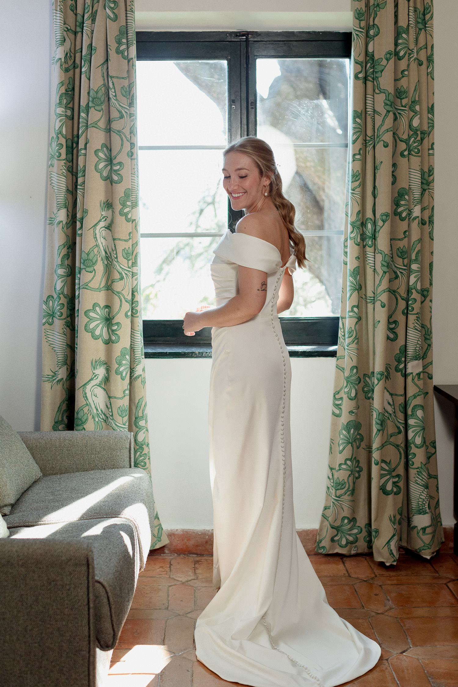 Una boda civil en el Parador de Jarandilla 14 La novia, de pie junto a una ventana con cortinas verdes estampadas, sonríe mientras luce su vestido blanco de hombros caídos y botones en la espalda, con la luz natural iluminando la escena.