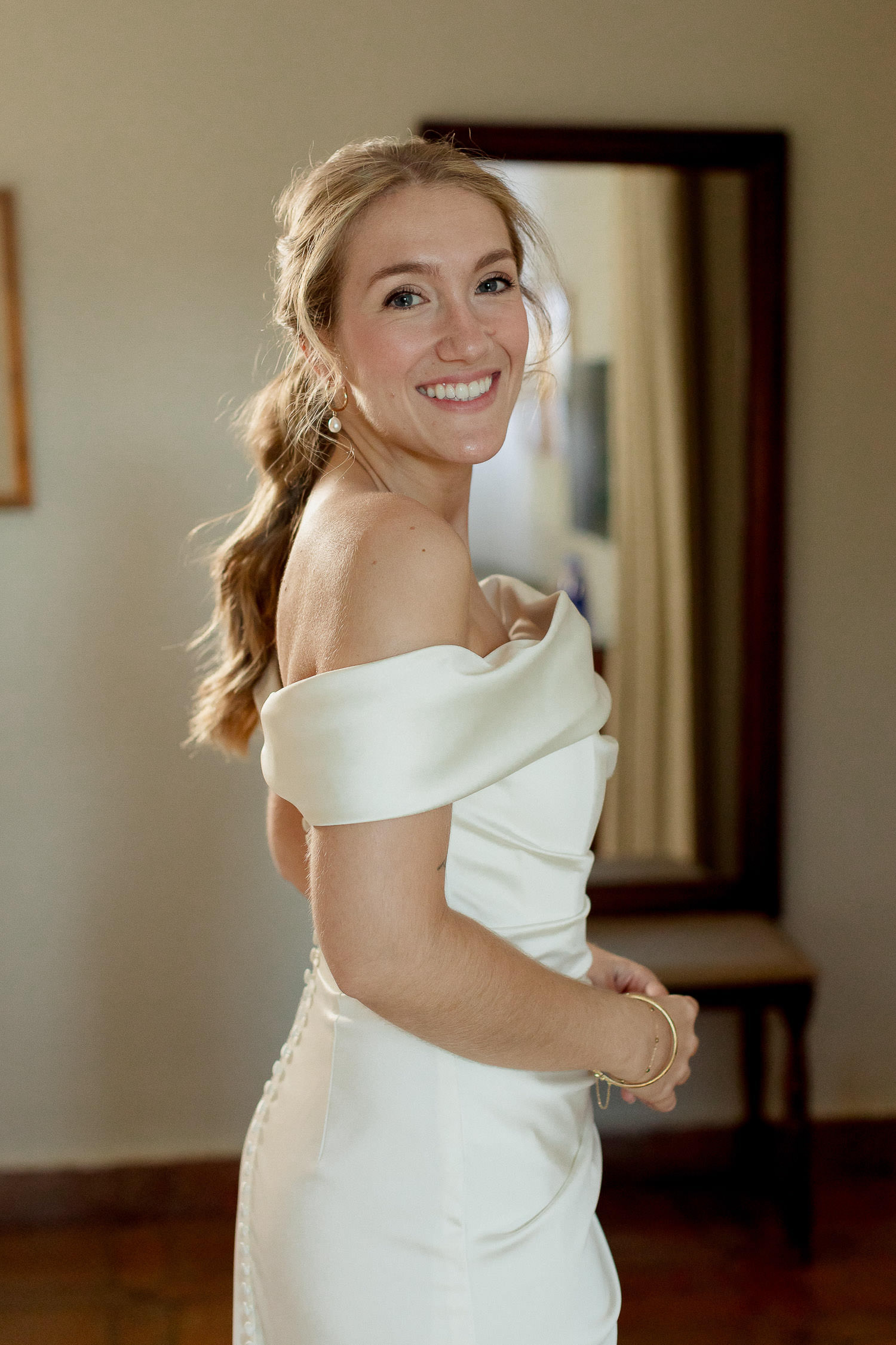Una boda civil en el Parador de Jarandilla 17 La novia sonríe ampliamente mientras posa de perfil, luciendo su vestido blanco de escote barco y una pulsera dorada, con la calidez de la habitación realzando su felicidad.