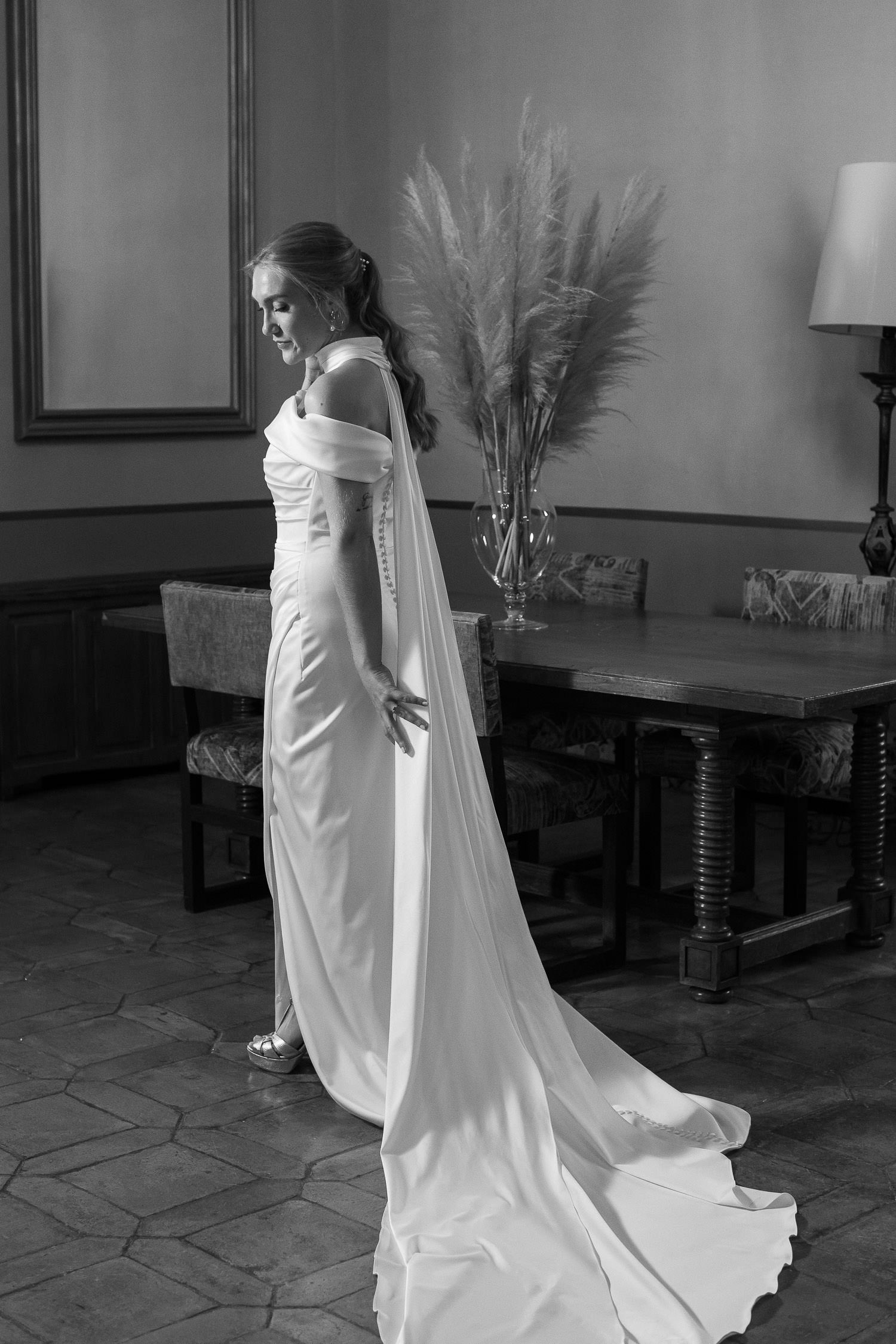 Una boda civil en el Parador de Jarandilla 24 La novia, radiante y llena de serenidad, luce un elegante vestido blanco de satén con capa larga y botones en la espalda, lista para su boda civil en el Parador de Jarandilla de la Vera. Las imágenes en blanco y negro resaltan la elegancia y el ambiente íntimo del lugar.