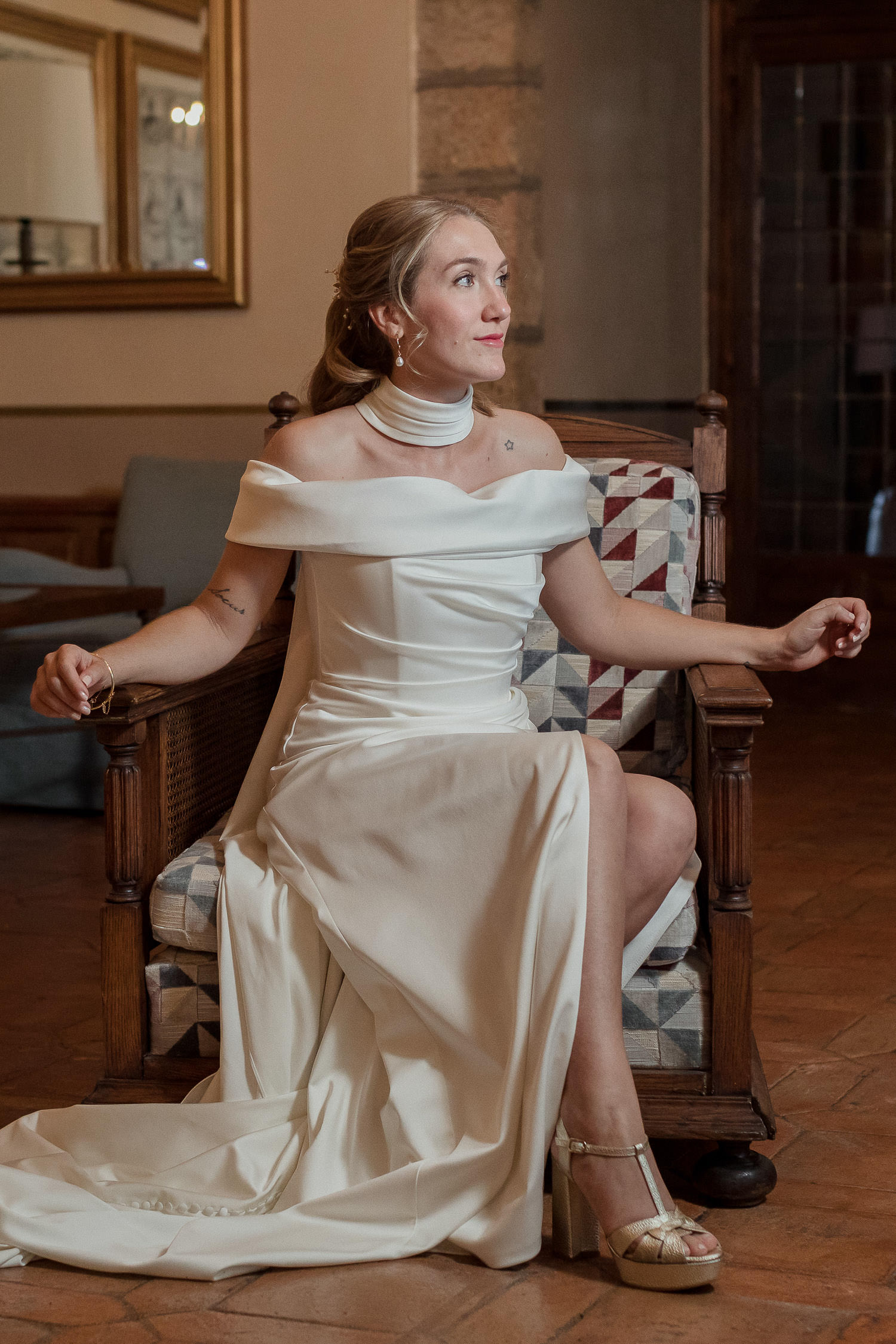 Una boda civil en el Parador de Jarandilla 26 La novia, sentada con elegancia en un sillón clásico de madera con cojines de patchwork en el Parador de Jarandilla, luce su vestido blanco con hombros descubiertos y un moderno cuello alto. La habitación, con paredes de piedra y suelo de baldosas, evoca la calidez y el encanto rústico del lugar.