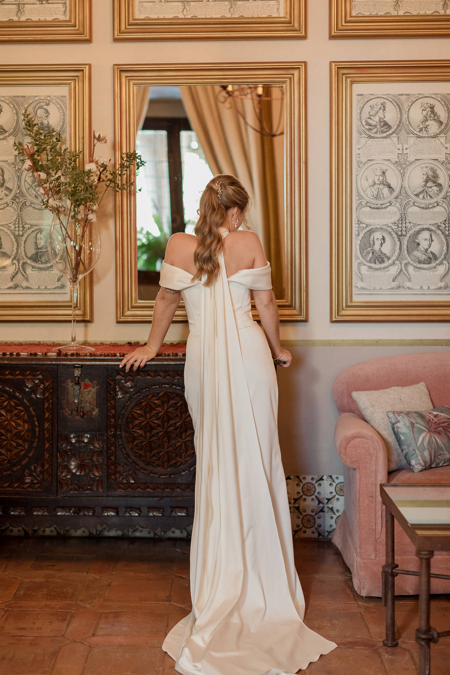 Una boda civil en el Parador de Jarandilla 27 Novia de espaldas luciendo su elegante vestido blanco de Eva Lendel en el Parador de Jarandilla. El ambiente combina el mobiliario clásico y toques naturales, con un espejo y cuadros antiguos dorados enmarcando la escena. Perfecto para una boda civil elegante y con historia.